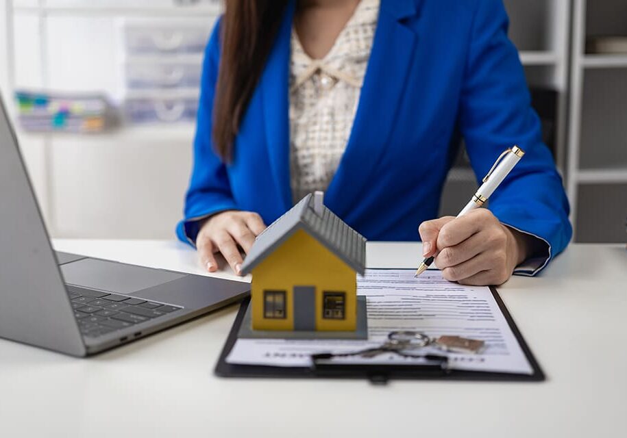 A person in a blue suit writes on a document beside a laptop. A small house model and a set of keys are on the desk.