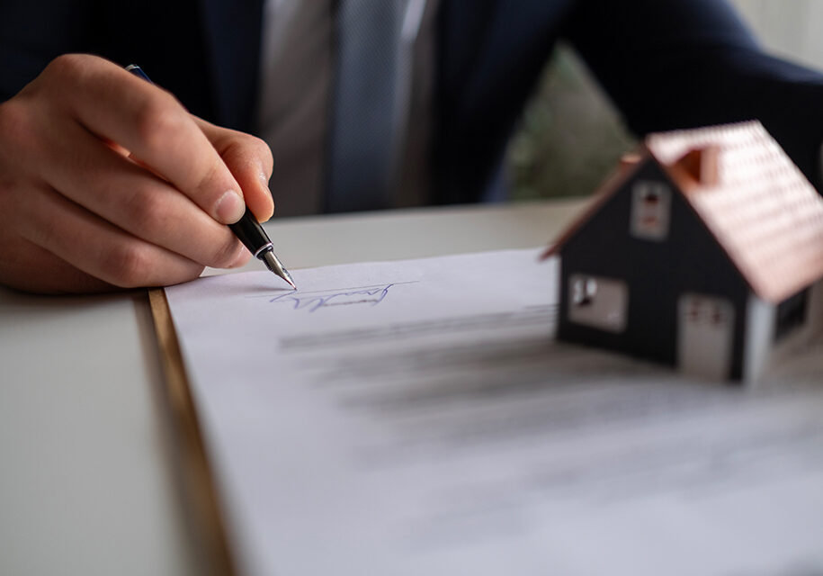 A person signs a document on a table beside a small model house, symbolizing a real estate transaction.